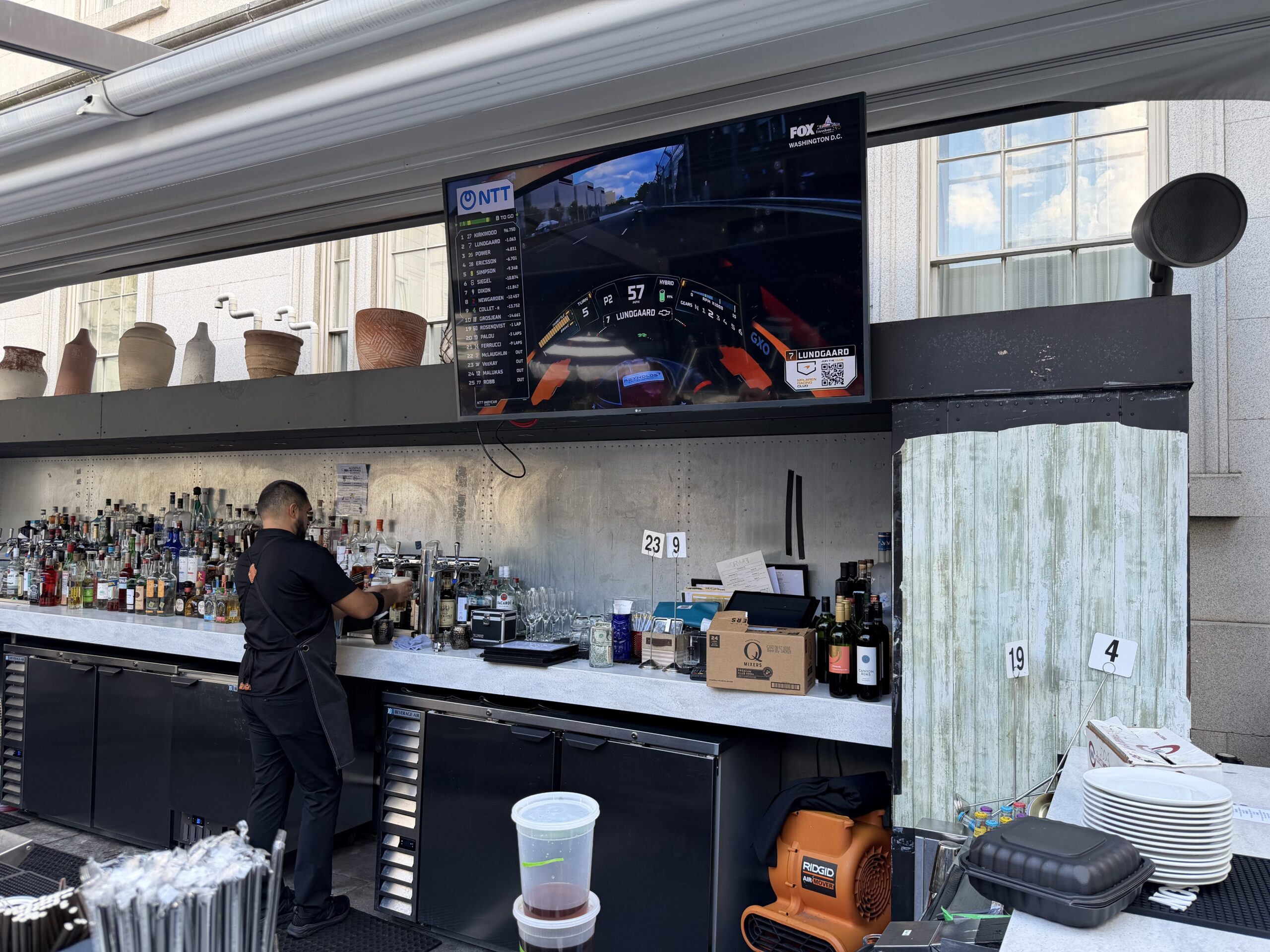 A bartender works behind an outdoor bar with various bottles, while a racing video plays on a large TV screen mounted above the counter.