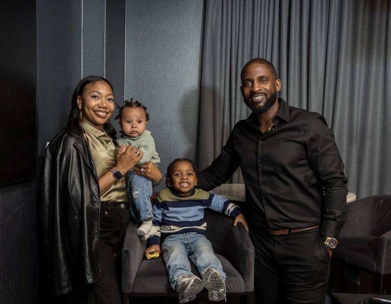A family of four poses indoors; a woman holds a baby, a man stands beside a child sitting on a chair. All are smiling and dressed in casual and semi-formal clothes.