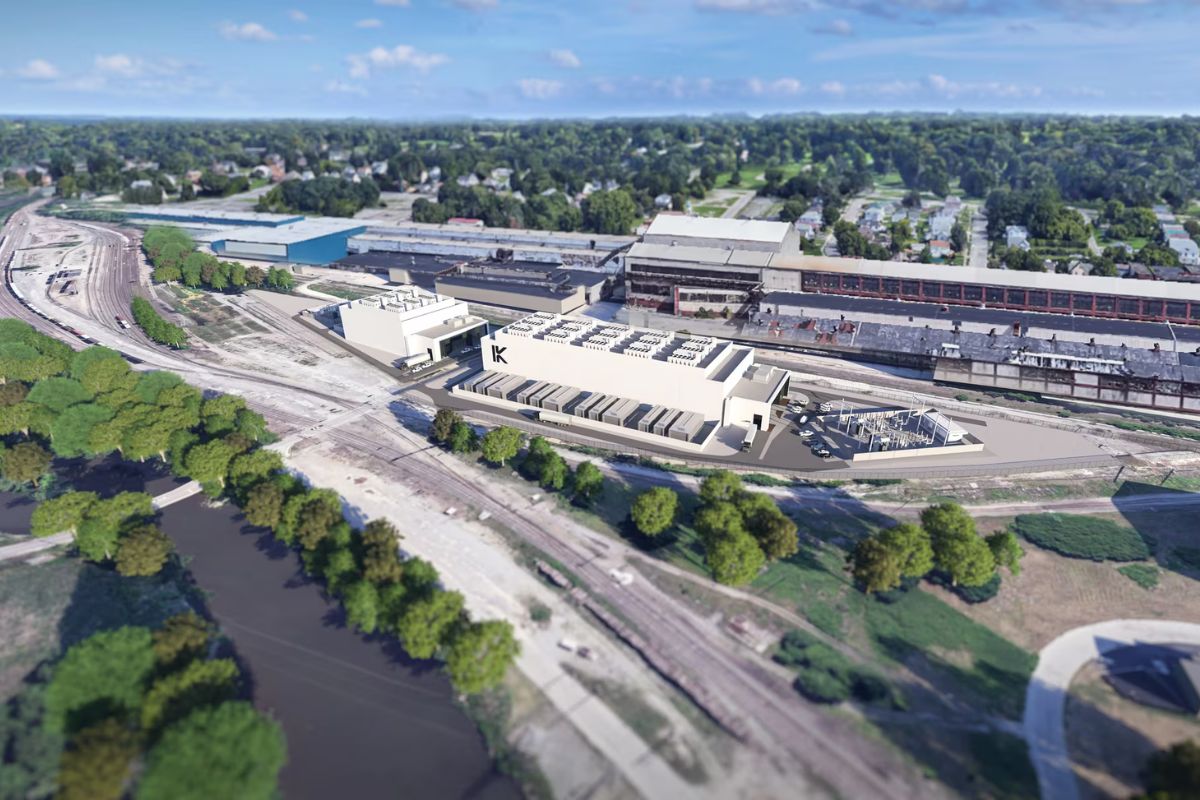 Aerial view of a large industrial facility near train tracks, surrounded by trees, with residential buildings and a river in the background.