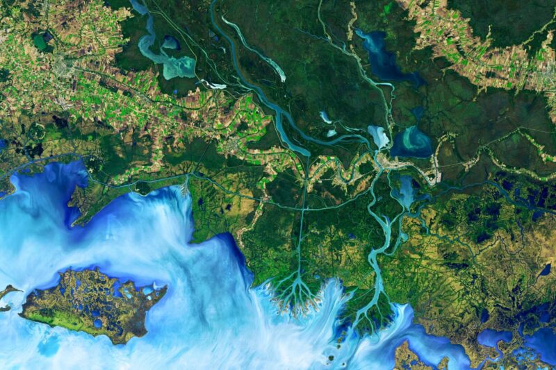Satellite image of a river delta with branching waterways meeting the sea, surrounded by green land, fields, and blue water.