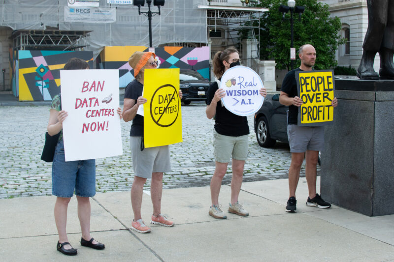 Four people stand outdoors holding protest signs about data centers and artificial intelligence, including messages like “Ban Data Centers Now!” and “People Over Profit.”.