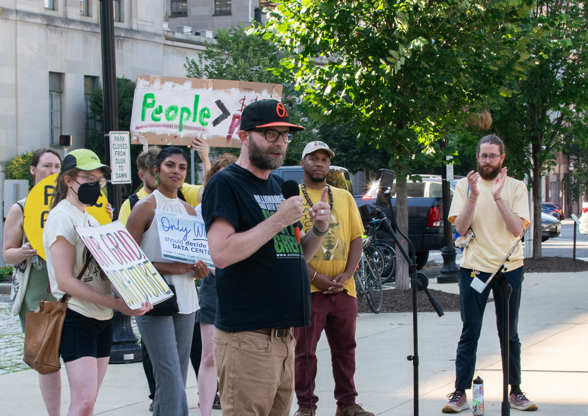 A person speaks into a microphone at an outdoor event. Several people stand nearby, some holding protest signs, and one person applauds. Trees and buildings are visible in the background.