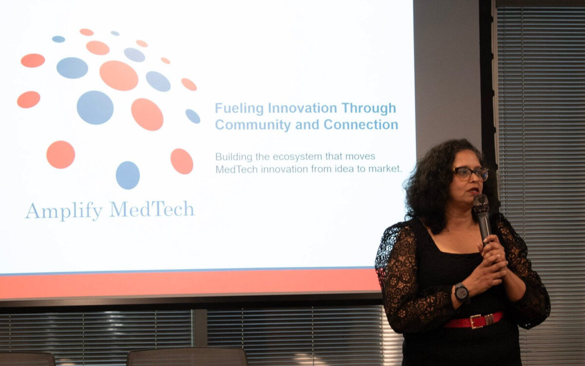 A woman speaks into a microphone in front of a screen displaying the Amplify MedTech logo and a slogan about innovation, community, and MedTech ecosystem growth.