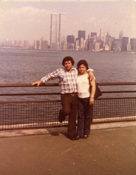A man and woman pose by a waterfront railing with the New York City skyline and the Twin Towers visible in the background.