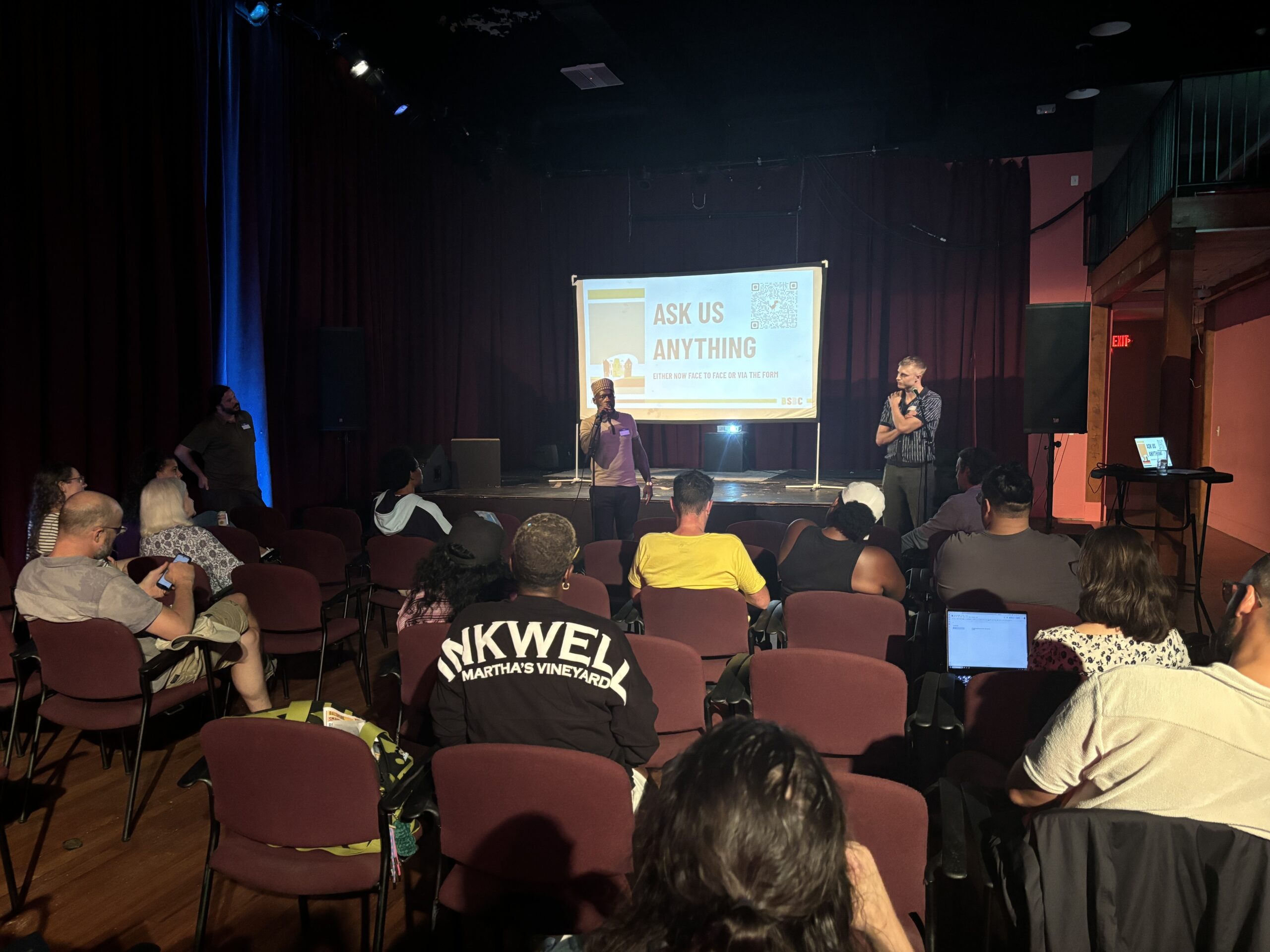 A group of people seated in an auditorium listen to two speakers standing at the front near a screen displaying “Ask Us Anything.”.
