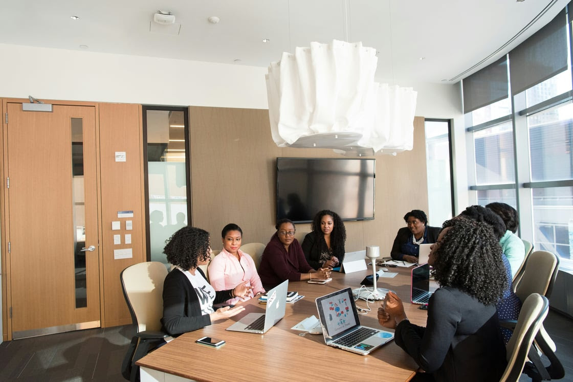 Nine people sit around a conference table with laptops and papers, engaged in a meeting inside a modern office with large windows and a wall-mounted TV.
