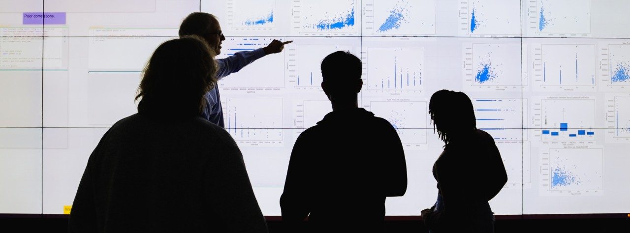 Four people observe a presenter pointing at graphs and scatter plots displayed on a large, brightly lit screen in a modern setting.