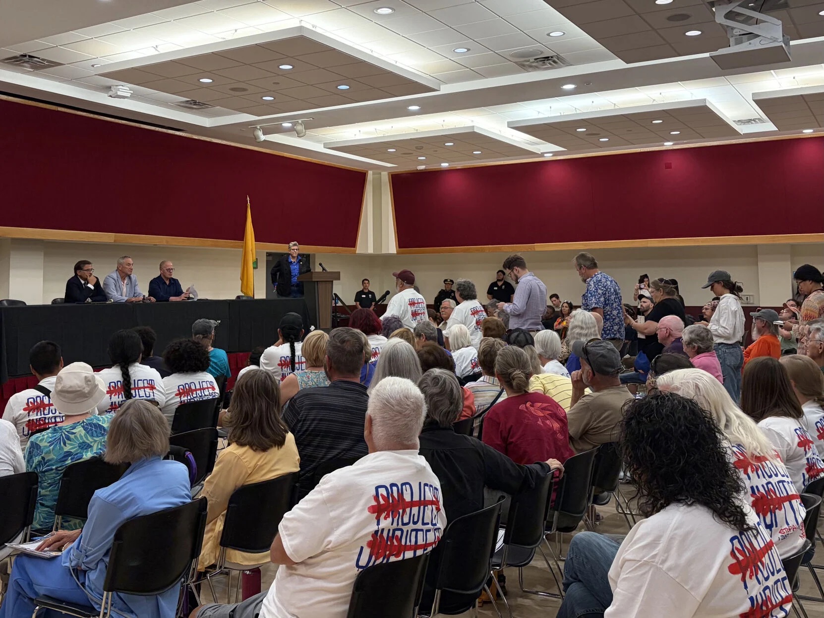 A large group of people attend a public meeting in a hall; some wear matching shirts reading “Protect Our Water.” A panel of speakers sits at a table, while one person speaks at the podium.