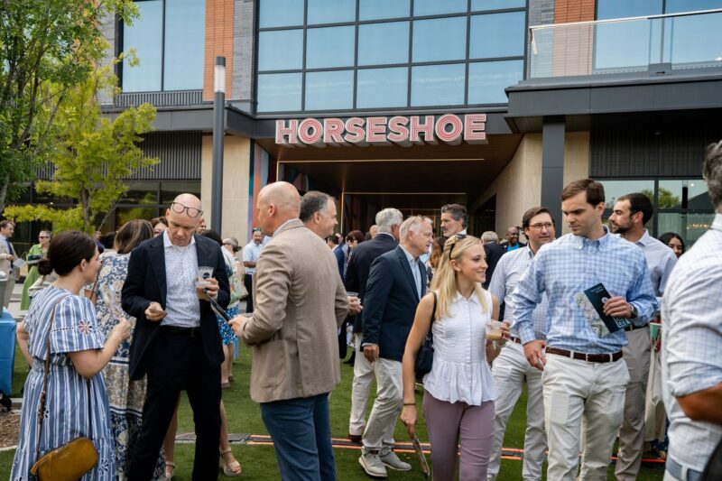 A group of people in business casual attire gather and socialize outside a building with a “HORSESHOE” sign.