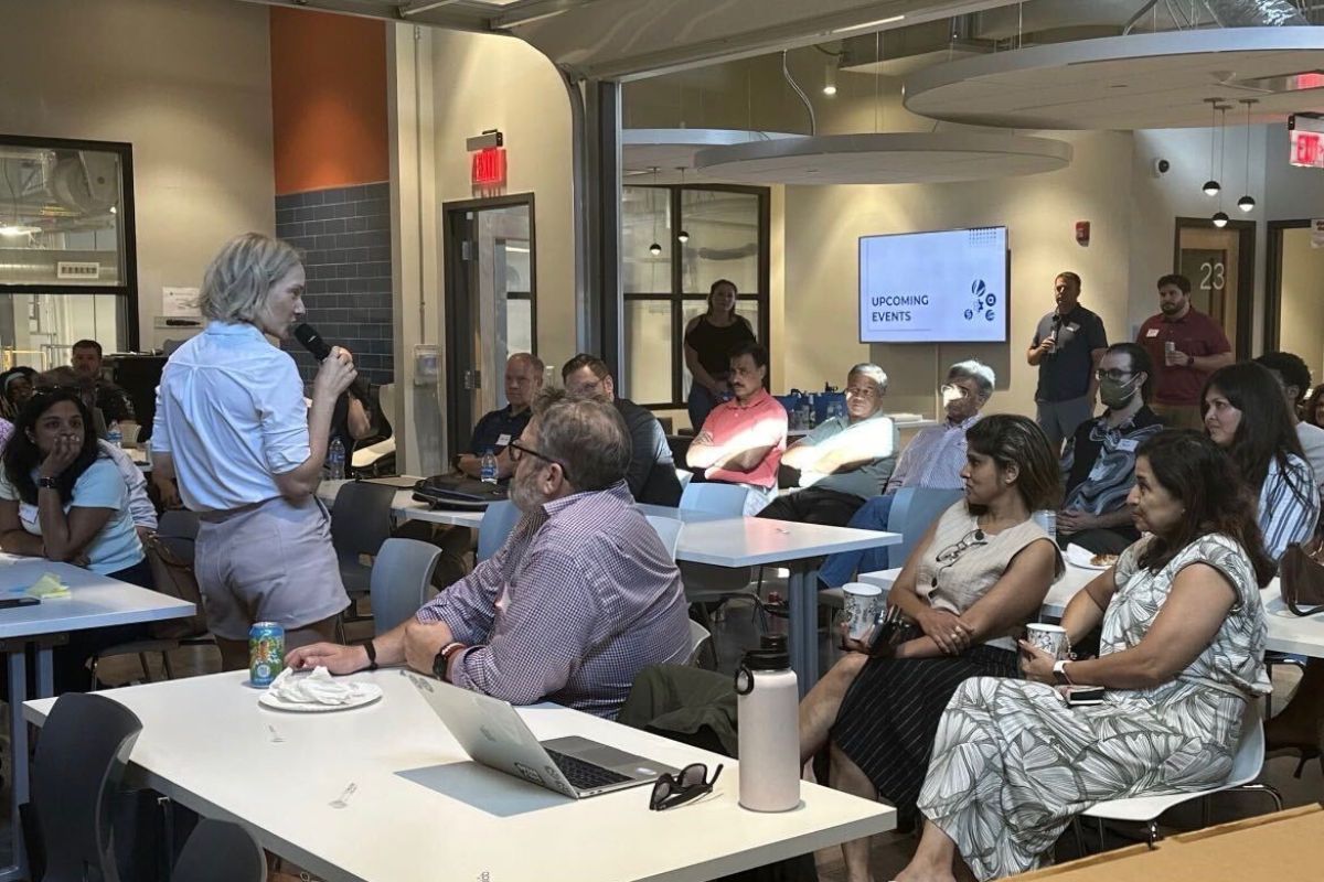 A woman speaks to a seated audience in a modern, well-lit meeting space. People listen attentively, some taking notes. A screen in the background displays “Upcoming Events.”.