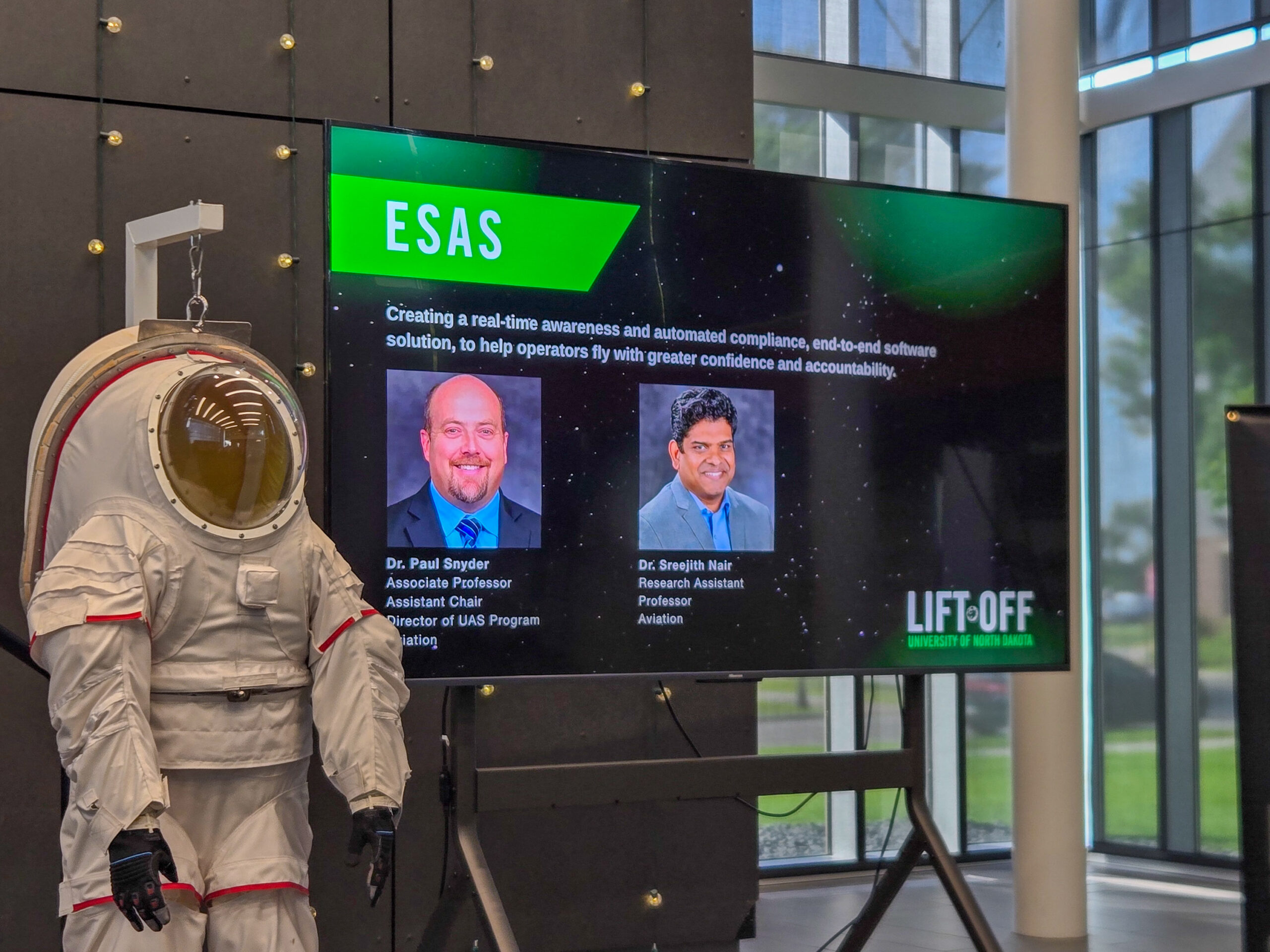 A mannequin in a spacesuit stands next to a screen displaying information about ESAS and two presenters at a LIFT-OFF event in a modern building.
