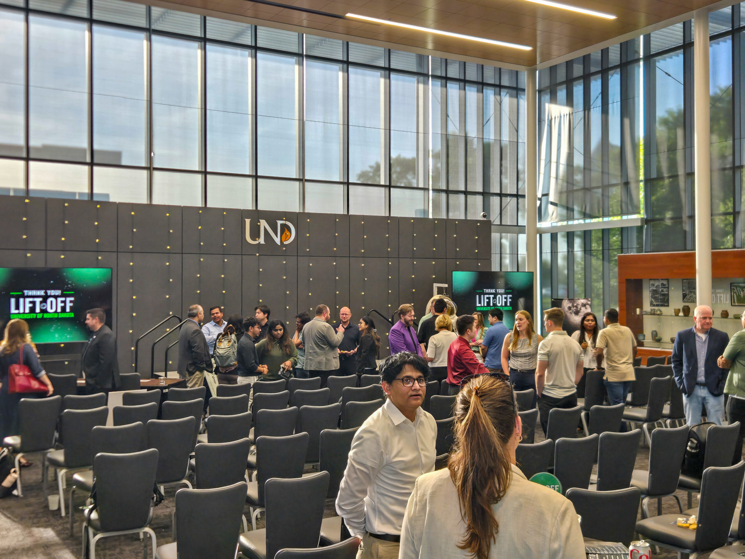 People gather and converse in a modern event space with rows of empty chairs, large windows, and a “Lift-Off” sign on the stage backdrop marked “UND.”.