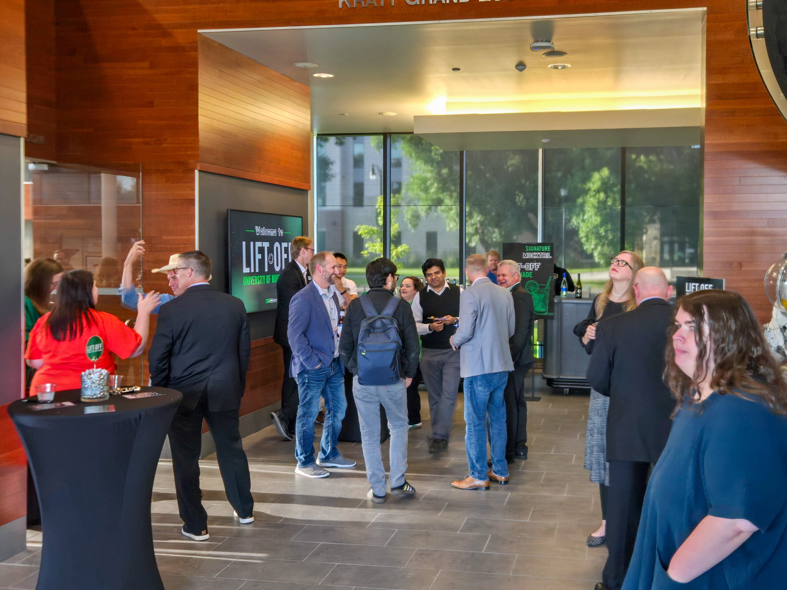 A group of people in business attire converses and networks in a modern lobby with wooden walls and large windows during a professional event.