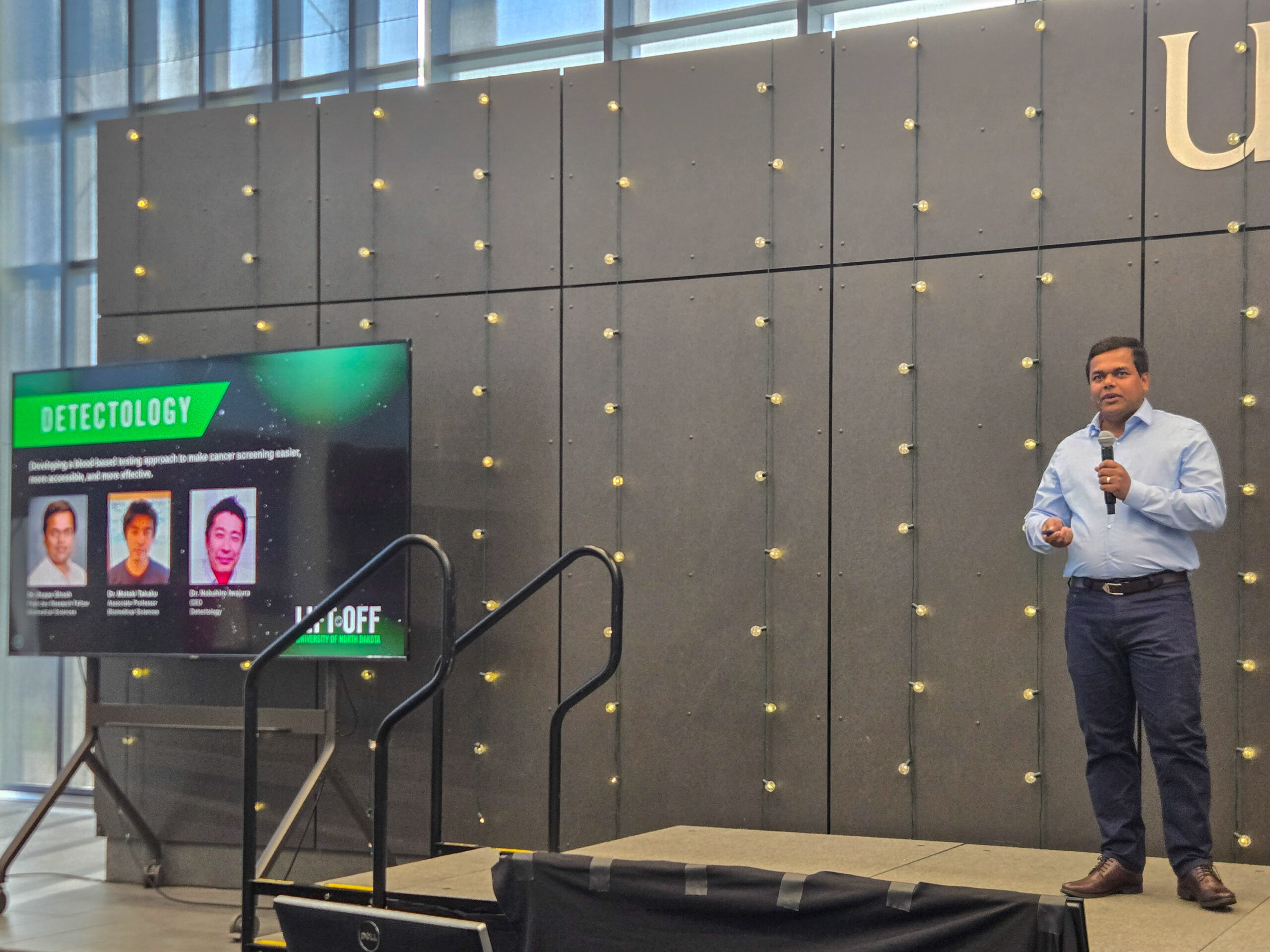 A man stands on stage giving a presentation next to a screen displaying information about “Detectology” and three pictured individuals.
