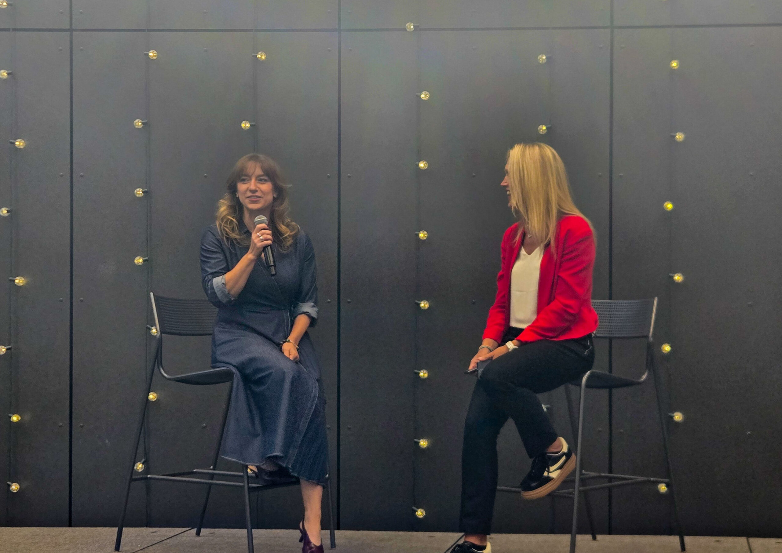 Two women sit on stools in front of a dark wall with small lights; one speaks into a microphone while the other listens.