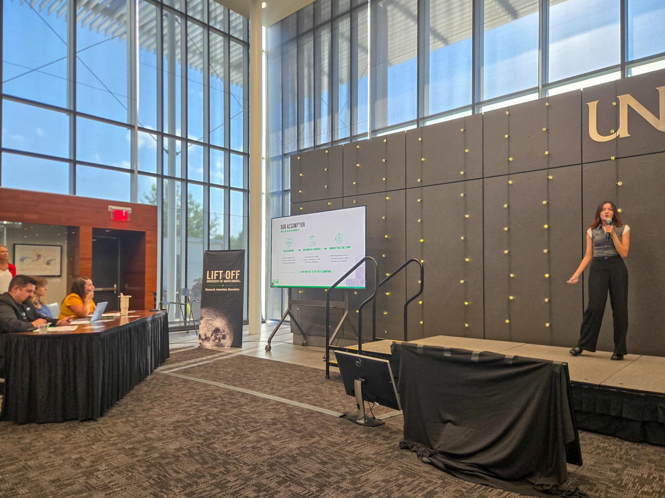 A woman is presenting a slideshow on stage to a panel of judges in a modern conference room with large windows and a “LIFT OFF” sign nearby.