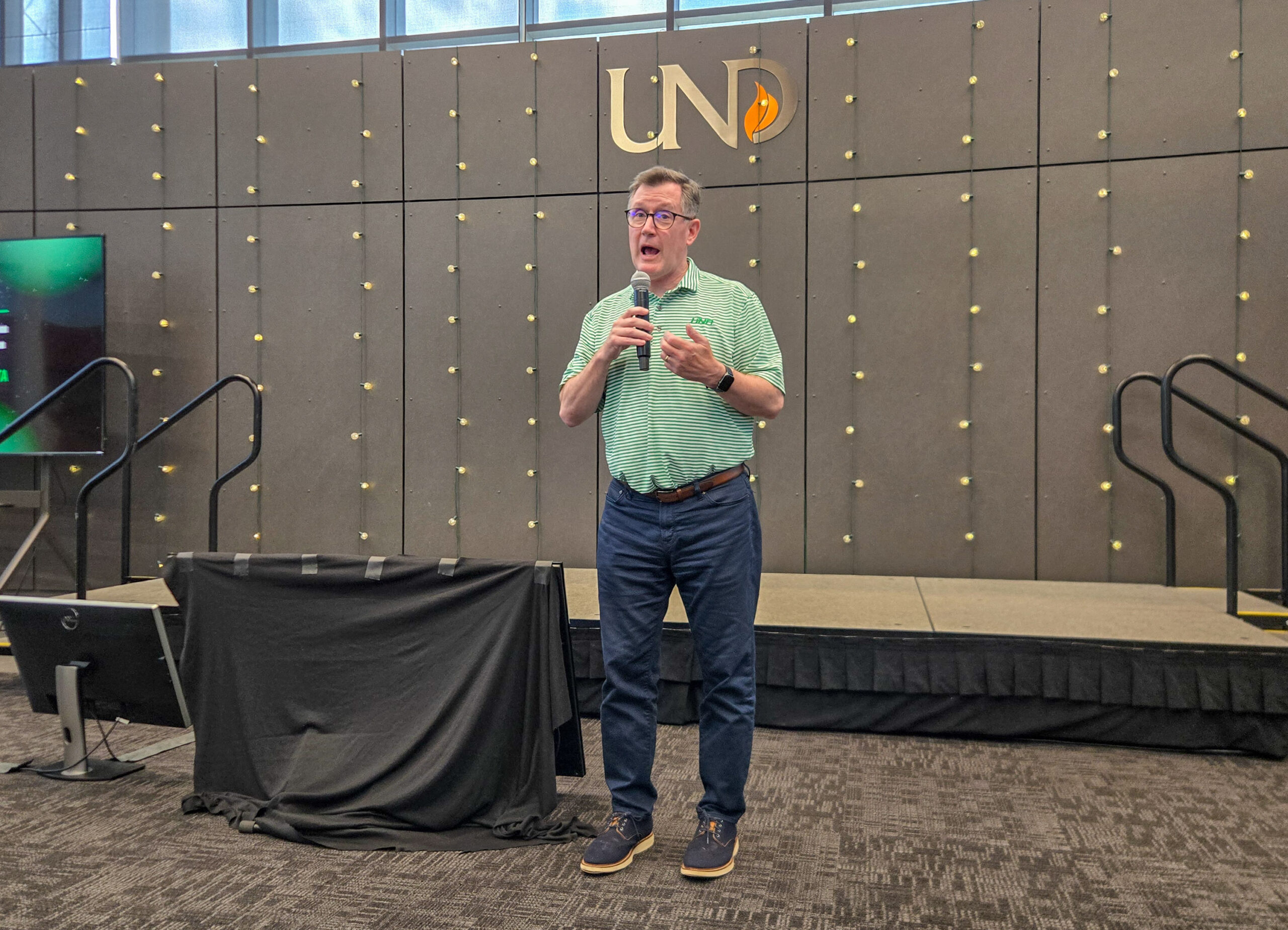 Man in a green shirt speaks into a microphone on a stage at a UND event, with a grey backdrop and string lights behind him.