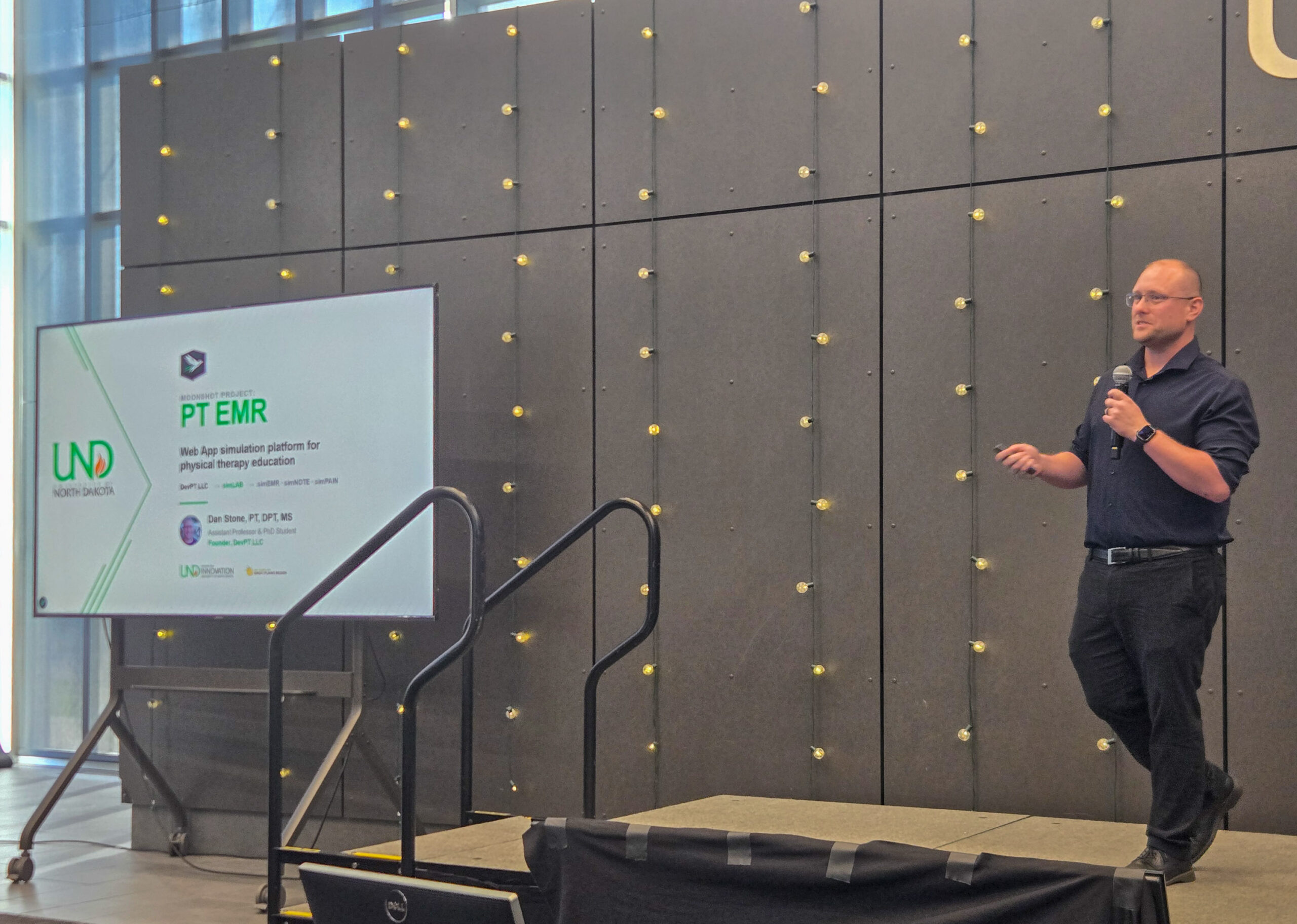 A man stands on stage holding a microphone, presenting a slide titled “PT EMR” next to a screen with University of North Dakota and partner logos.