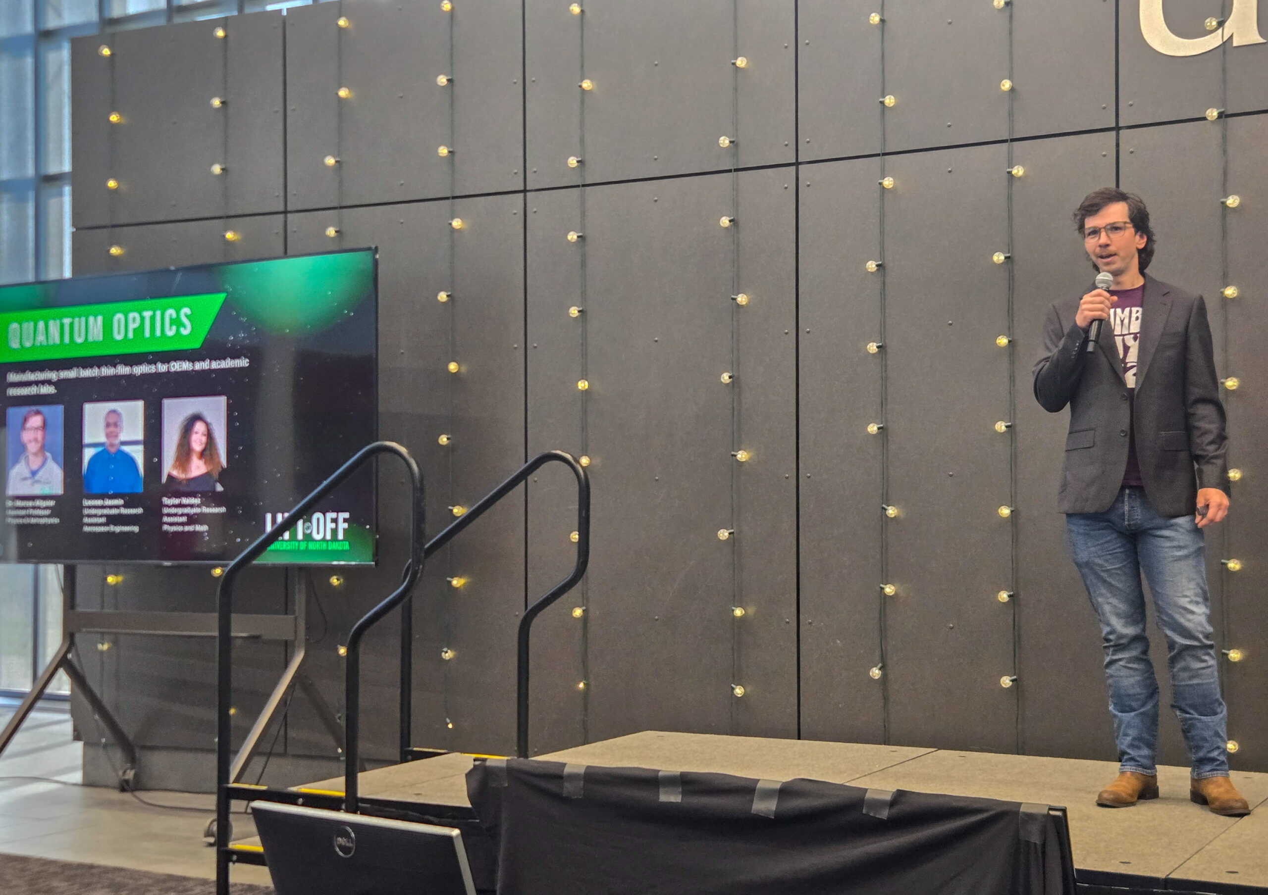 A man stands on stage holding a microphone beside a screen displaying a “Quantum Optics” panel with headshots and names of three people.