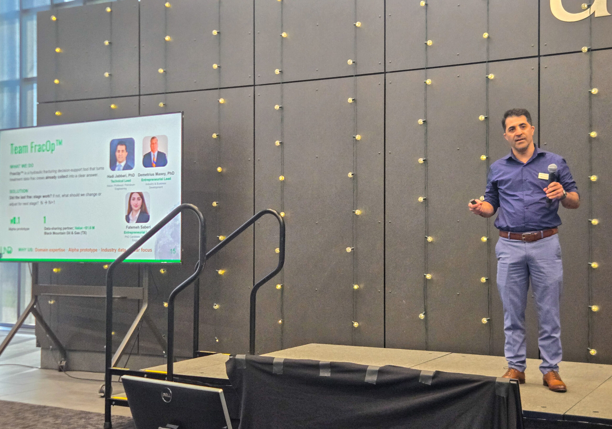 A man stands on a small stage giving a presentation; a screen displays information about a team with text and headshots.