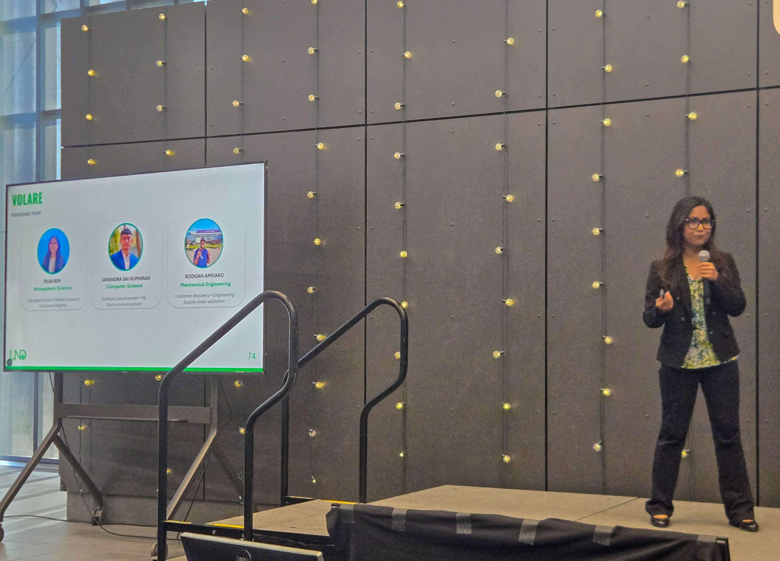 A woman in business attire gives a presentation on stage next to a screen displaying three profiles under the title “VOLARE.”.