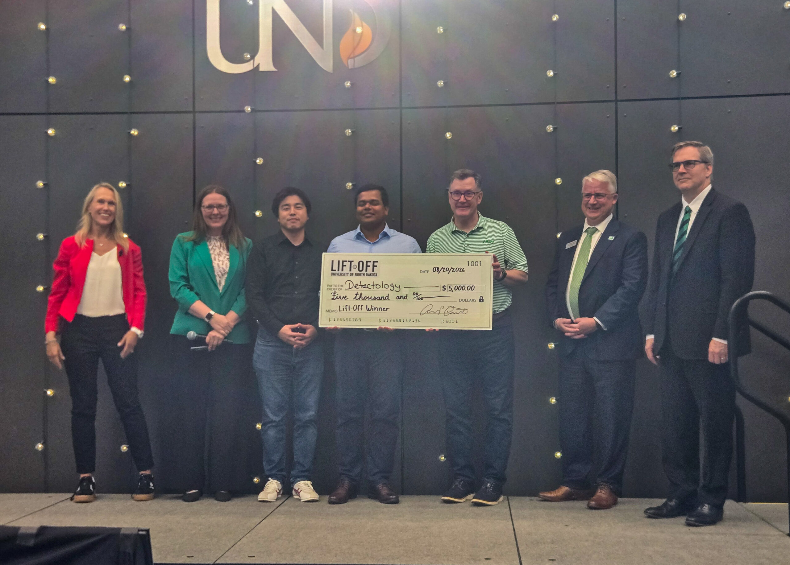 Six people stand on stage; four hold a large check labeled “LIFT-OFF” for $20,000, awarded to “Omniology” as the Lift Off Winner. The background features a dark wall with the “UND” logo.
