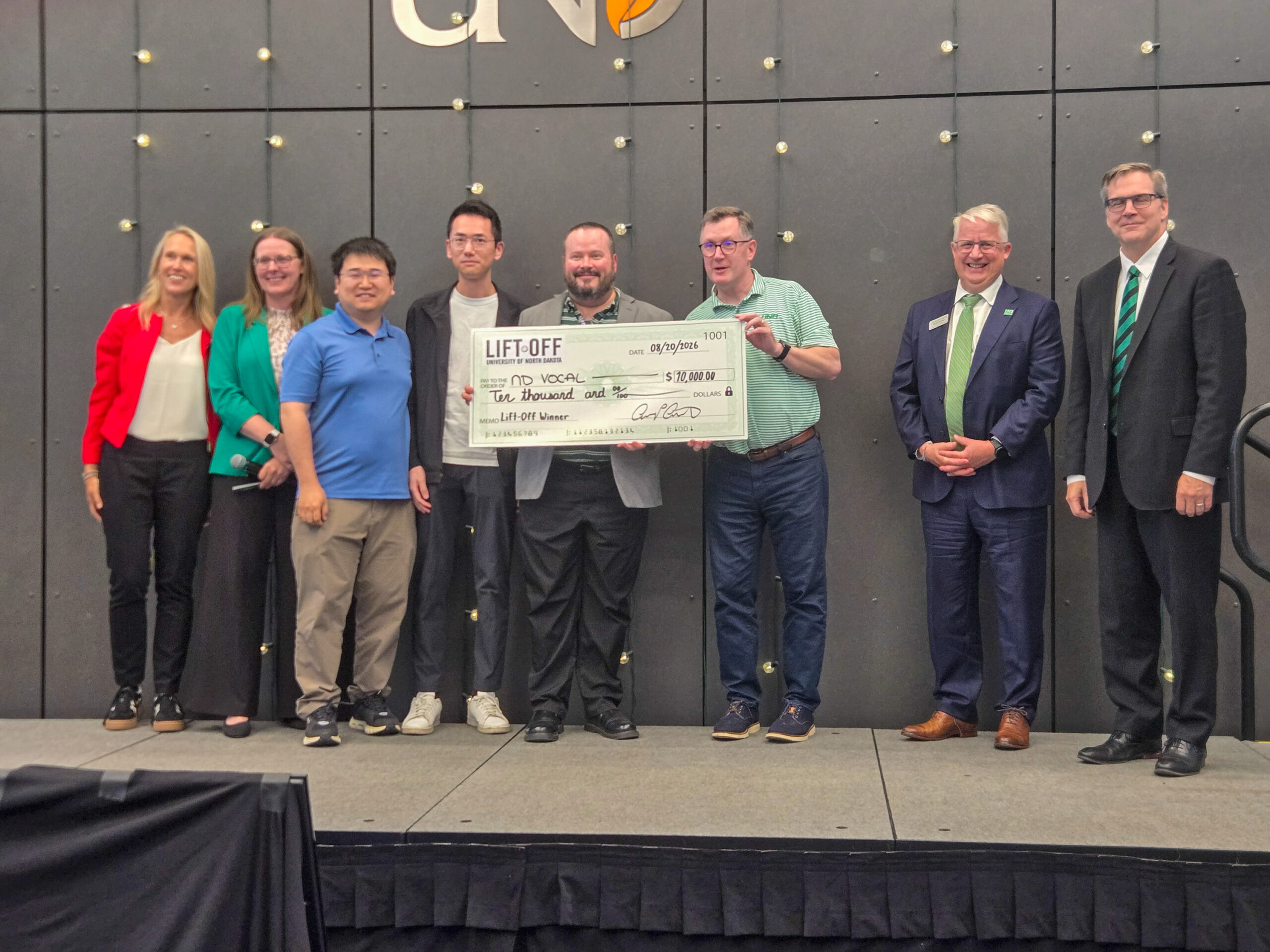 Seven people stand on a stage, posing with an oversized check for $10,000 labeled “LIFT-OFF” against a gray backdrop with the letters “CN” visible.