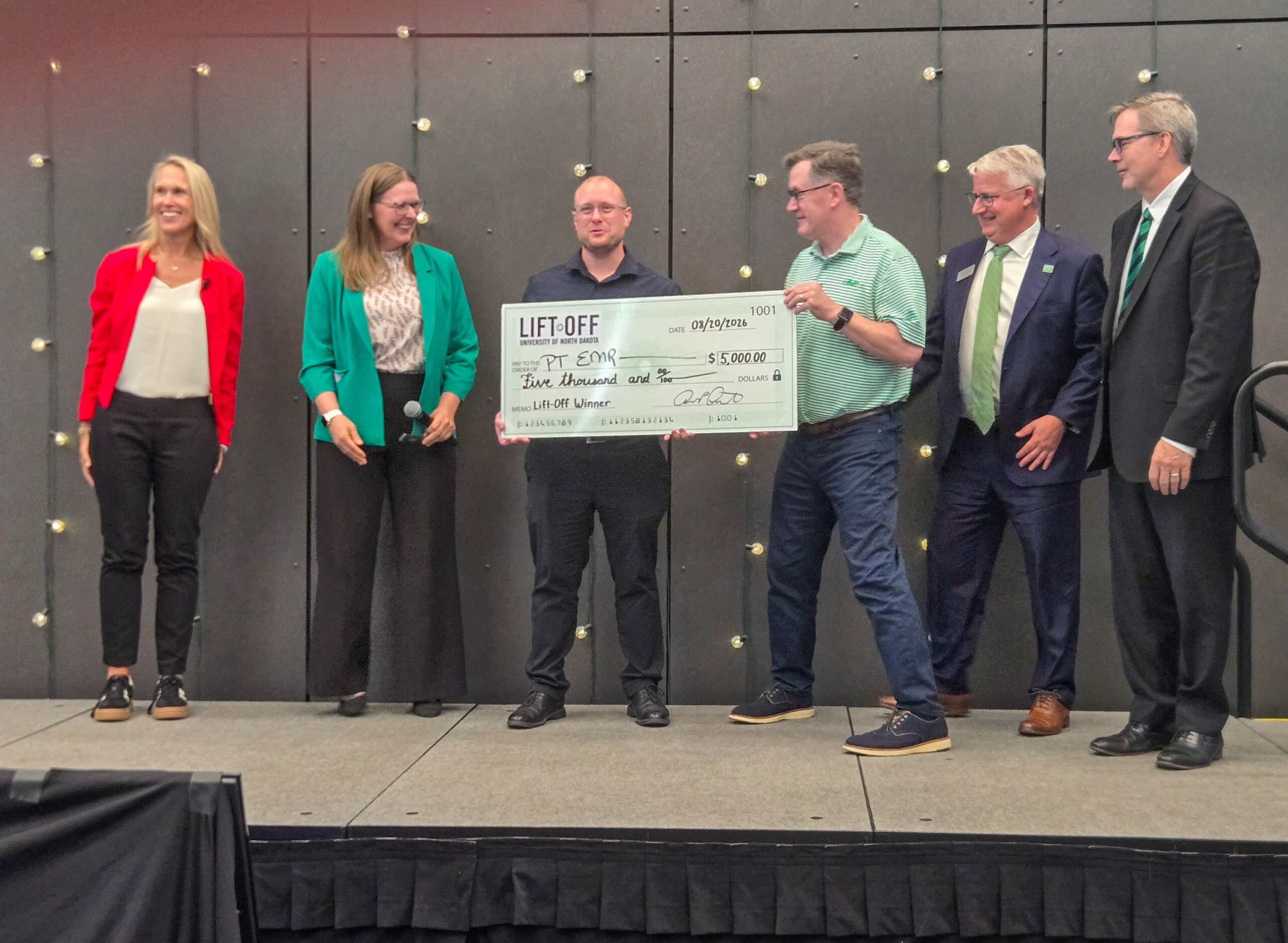 Six people stand on a stage; one holds a large ceremonial check for $3,000 made out to PT Genie from Lift Off.