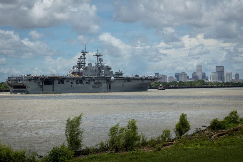 A large gray naval ship sails on a river with a city skyline and cloudy sky in the background; green foliage lines the riverbank.