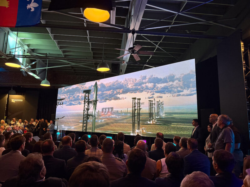 A crowd watches a presentation showing a rocket launch site on a large screen inside an indoor venue with overhead lighting and ceiling beams.