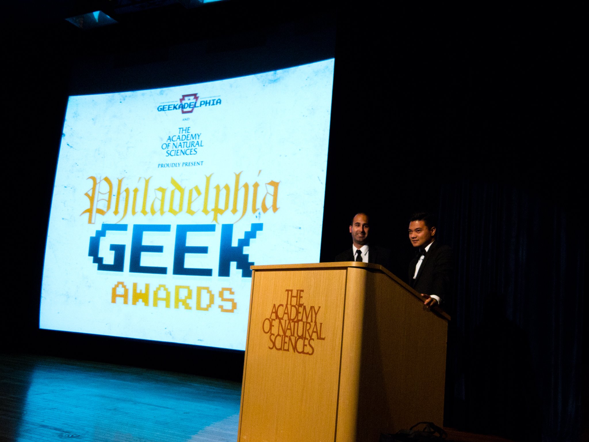 Two men in suits stand at a podium on stage with a large screen behind them displaying “Philadelphia Geek Awards” at The Academy of Natural Sciences.