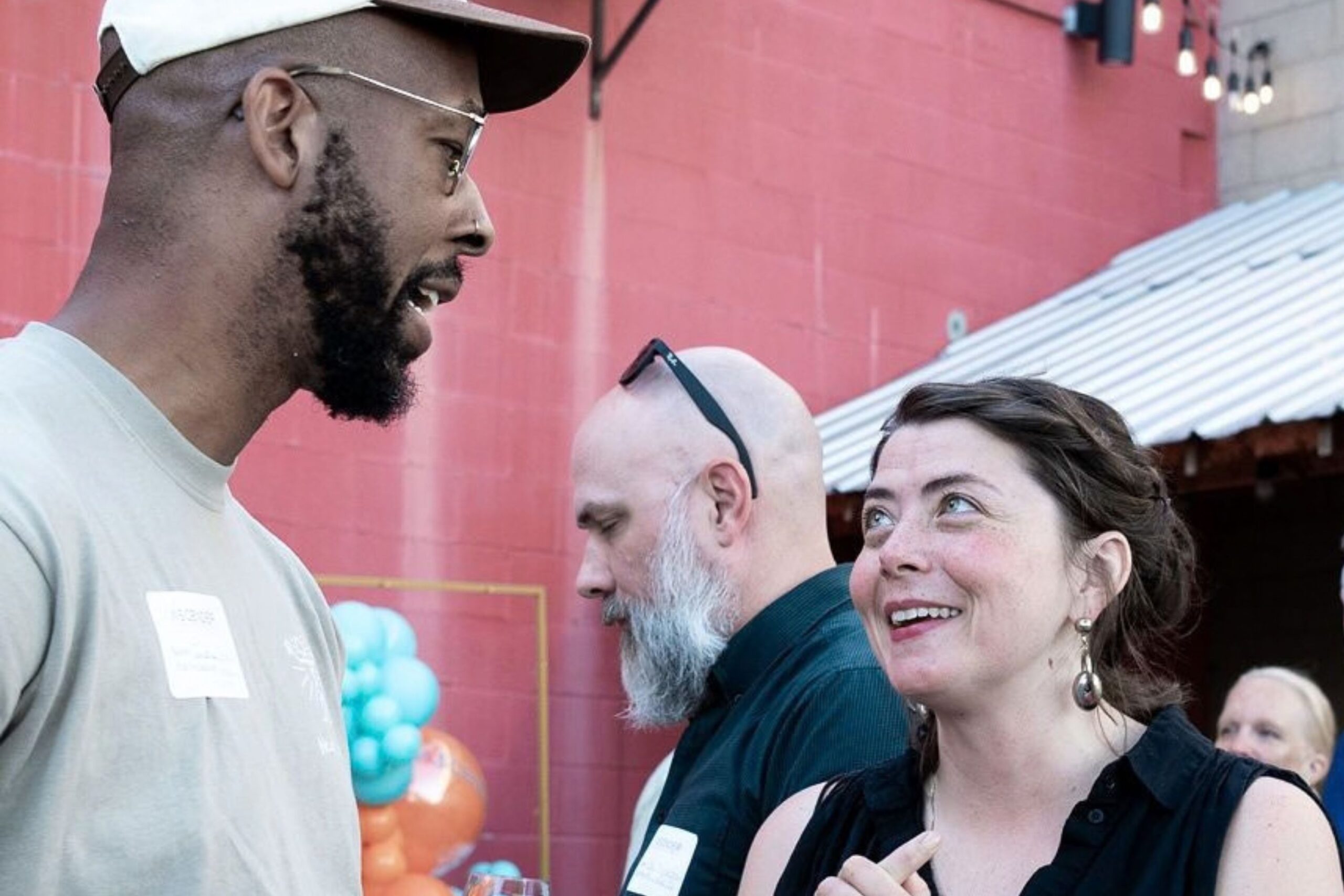 Three adults converse at an outdoor event; one man speaks while a woman smiles, and another man stands in the background. Balloons and a red wall are visible.