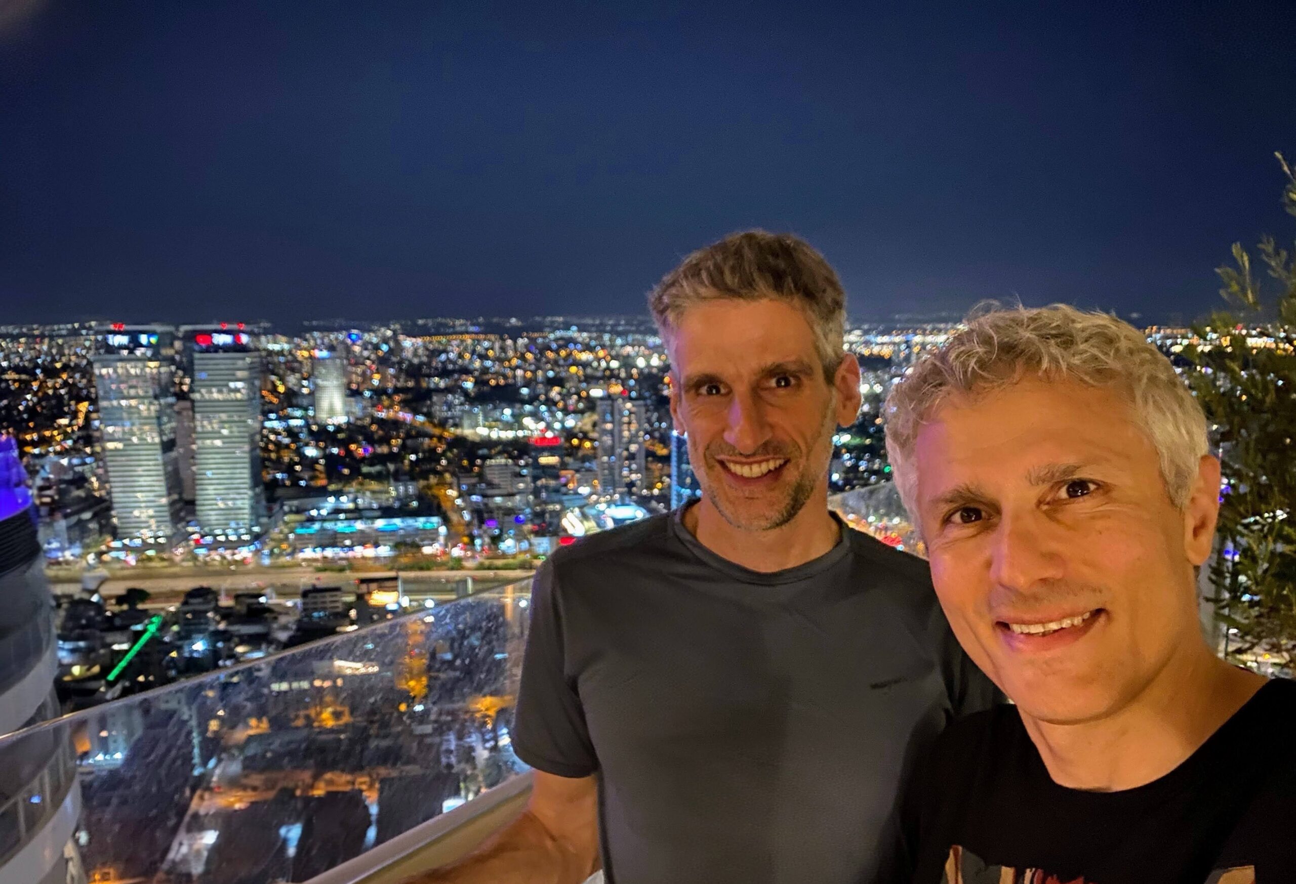 Two men stand on a balcony at night overlooking a brightly lit city skyline with tall buildings and many lights in the distance.