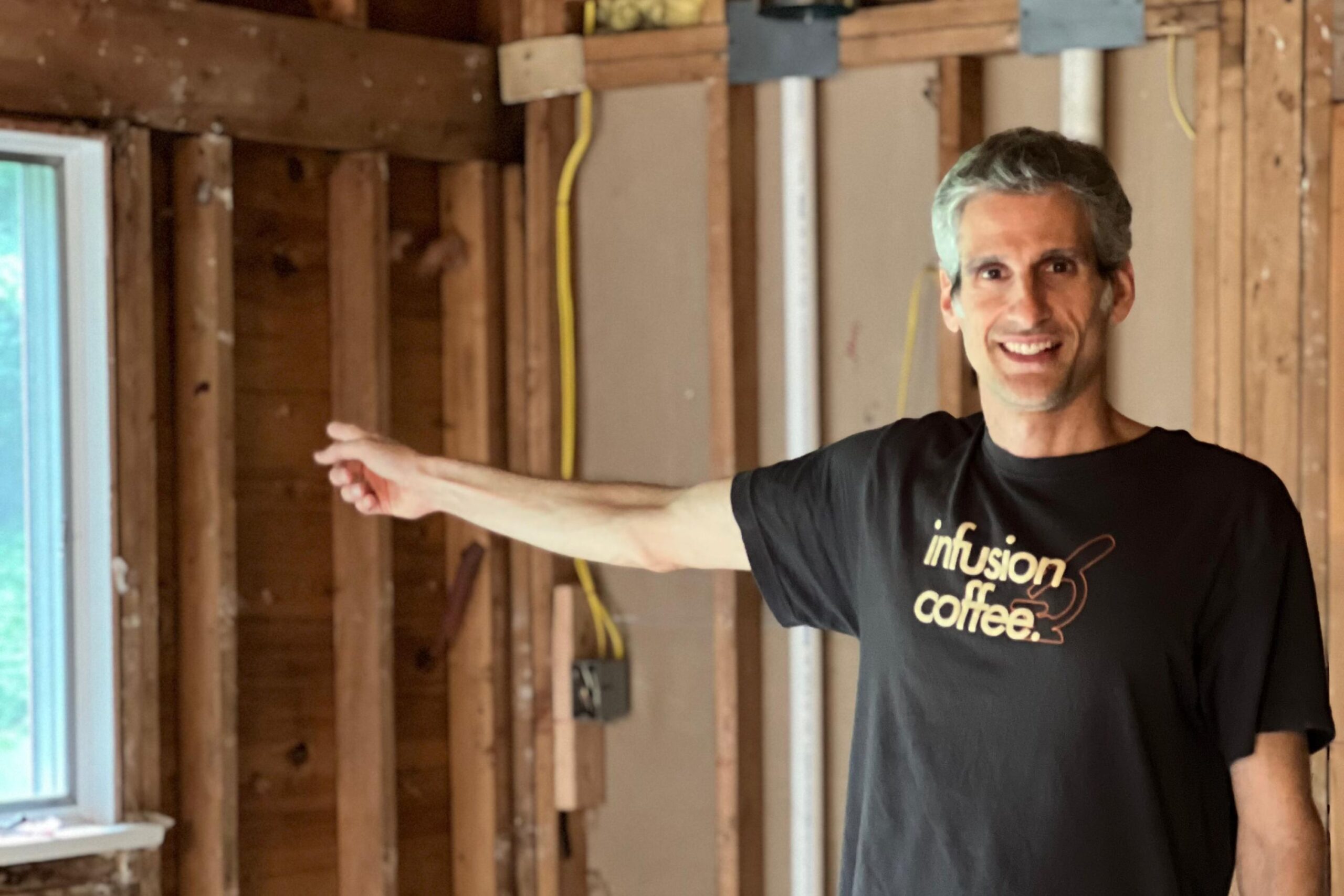 A man in a black “infusion of coffee” shirt stands in a partially constructed room, smiling and pointing toward exposed wooden wall studs and pipes.