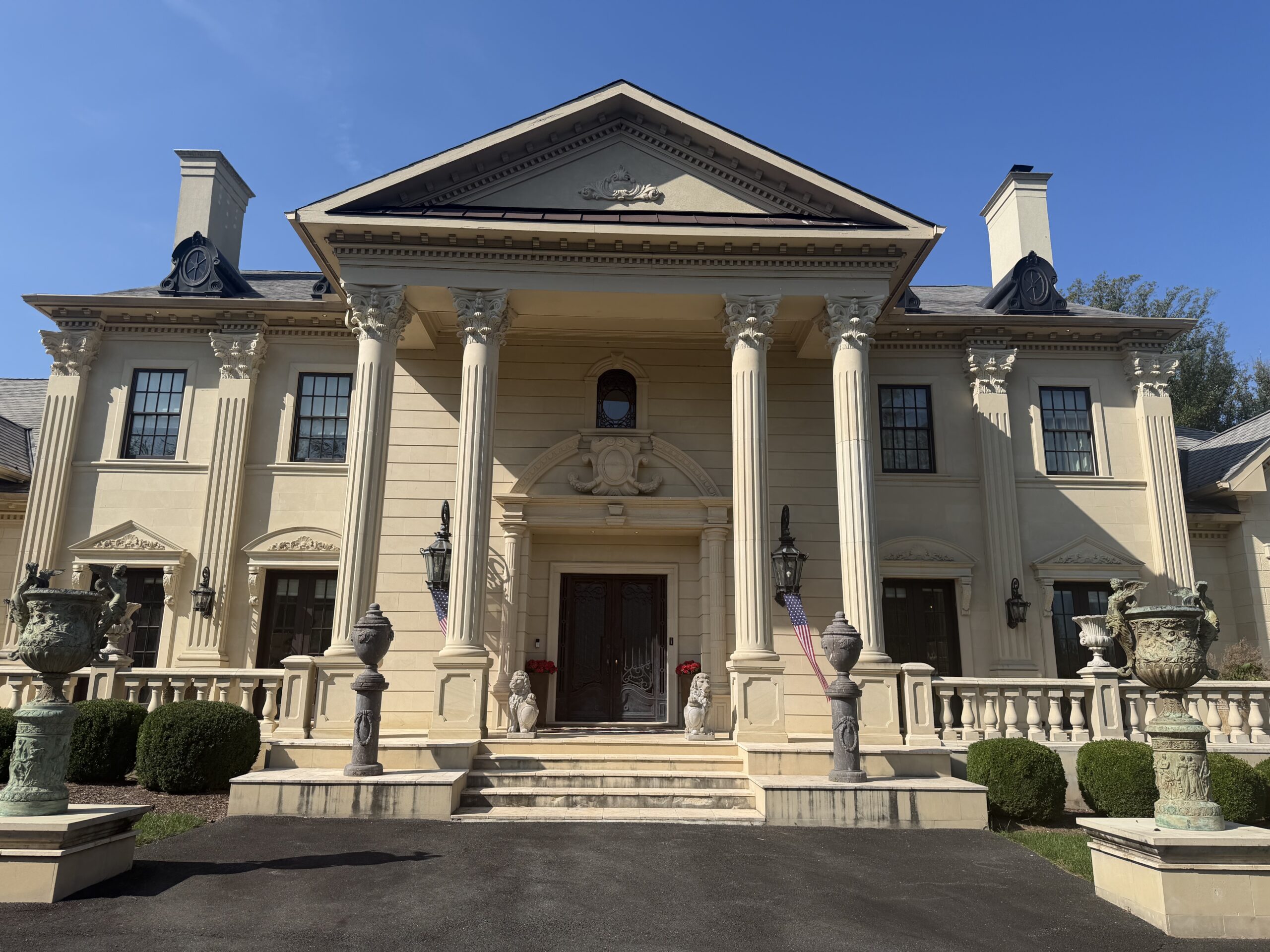 Large beige mansion with tall columns, ornate details, double front doors, stone urns, and manicured landscaping under a clear blue sky.