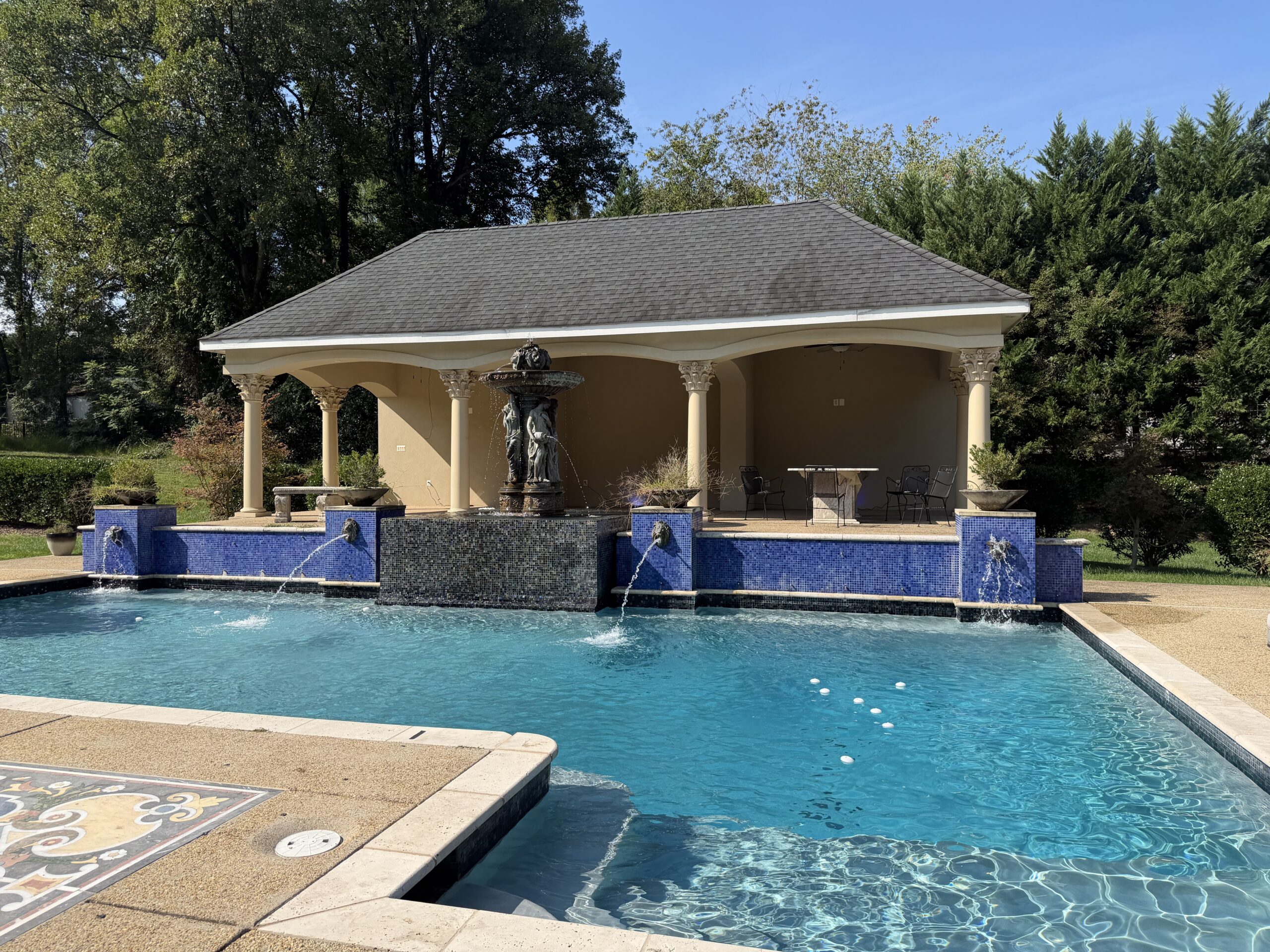 A rectangular swimming pool with a fountain and water features sits in front of a covered patio area with columns and outdoor furniture.