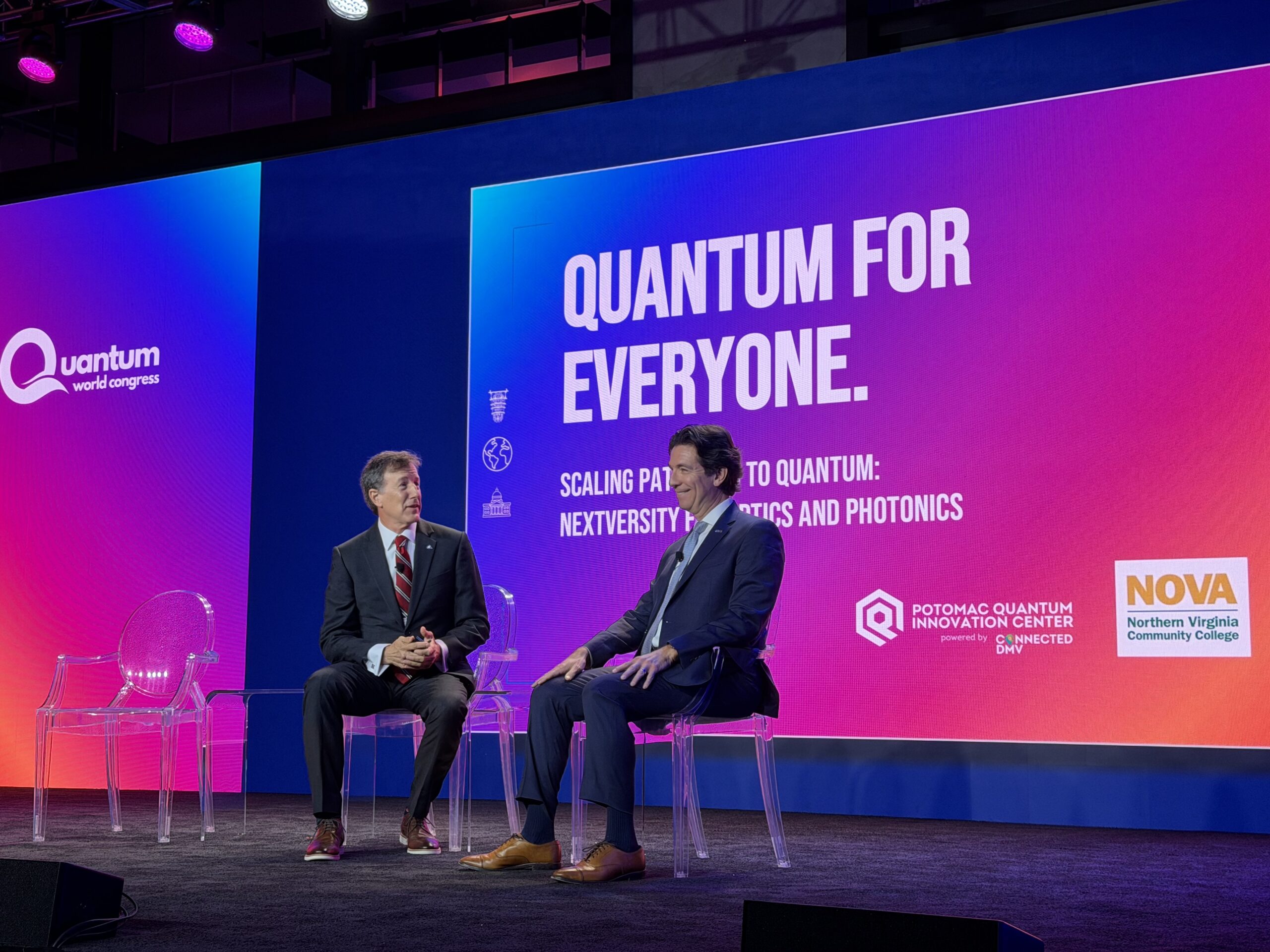 Two men in suits sit on stage discussing quantum technology, with a large screen behind them displaying “Quantum for Everyone” at a technology conference.