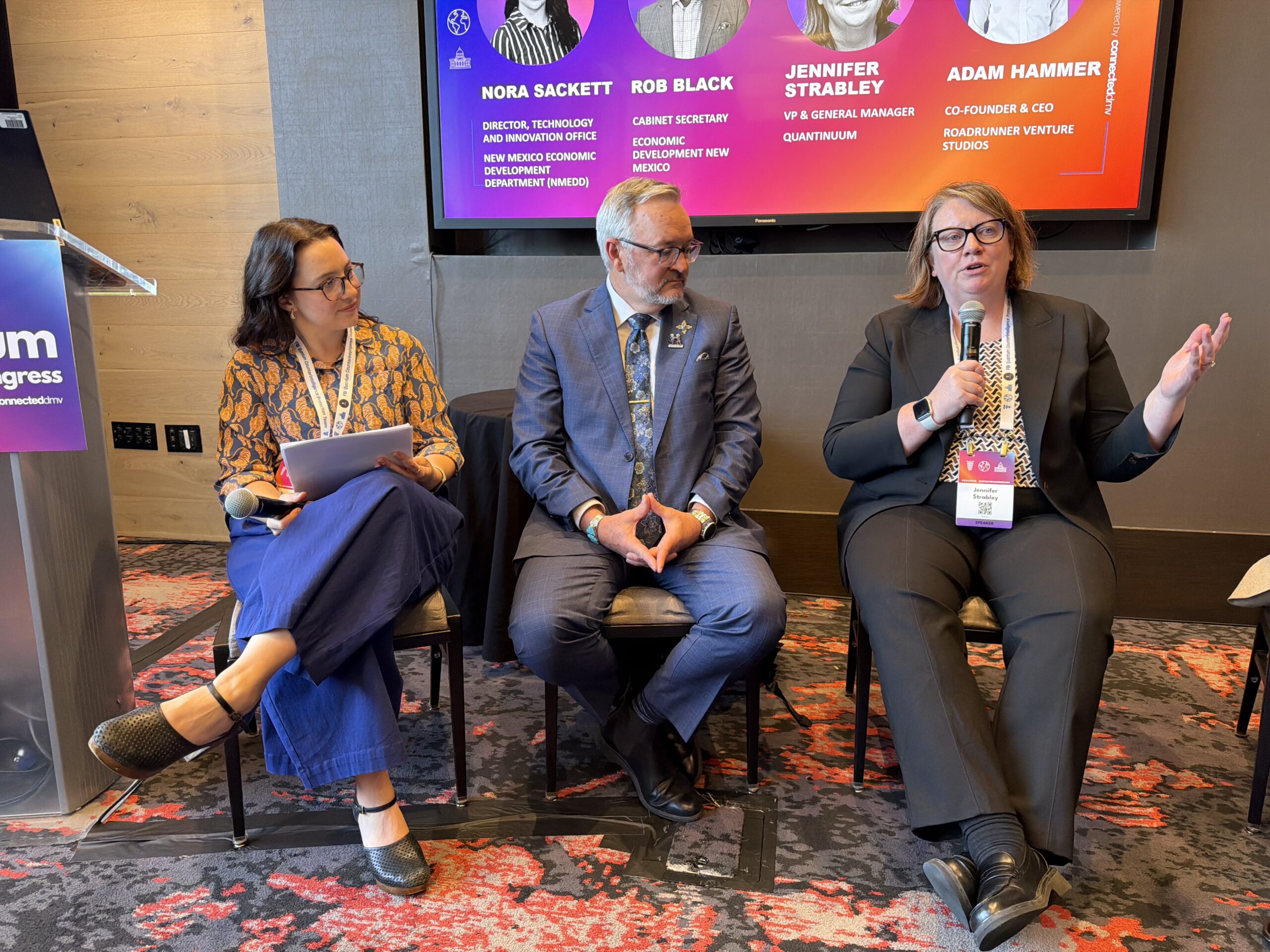 Three panelists sit on chairs at a conference, one woman speaks into a microphone while the others listen, with event name tags visible and a presentation slide in the background.