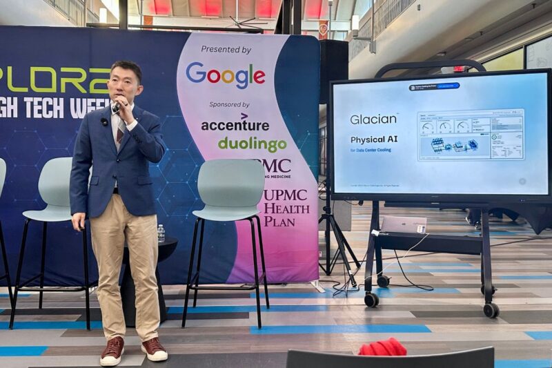 A man in a blue blazer speaks into a microphone next to a screen displaying a Glacian Physical AI presentation at a tech event.