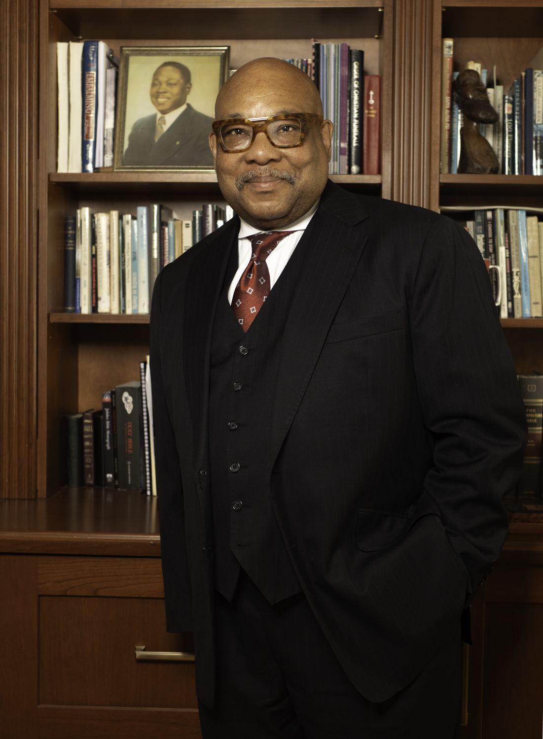 A man in a dark suit stands in front of a bookshelf filled with books and framed photos, looking at the camera with one hand in his pocket.