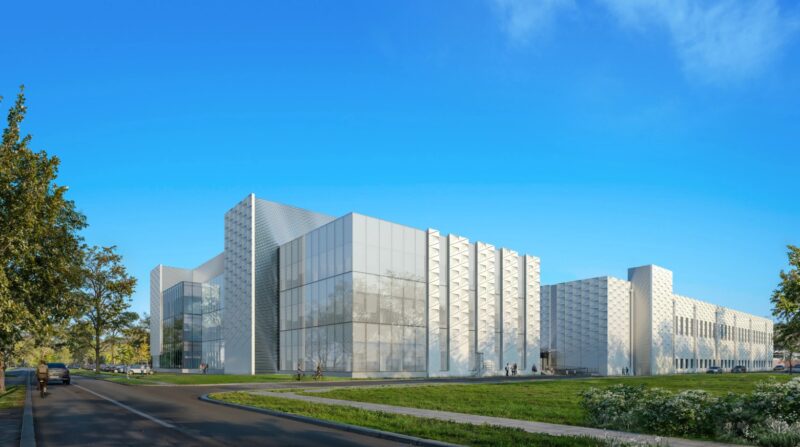 Modern white building with geometric facade panels, large windows, and surrounding green lawn under clear blue sky. A road runs by the front, with trees and people nearby.