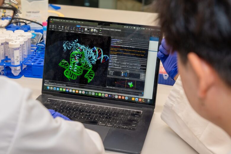 Two people in lab coats and gloves look at a laptop displaying a colorful 3D molecular model, with laboratory tubes and racks visible beside them on the table.