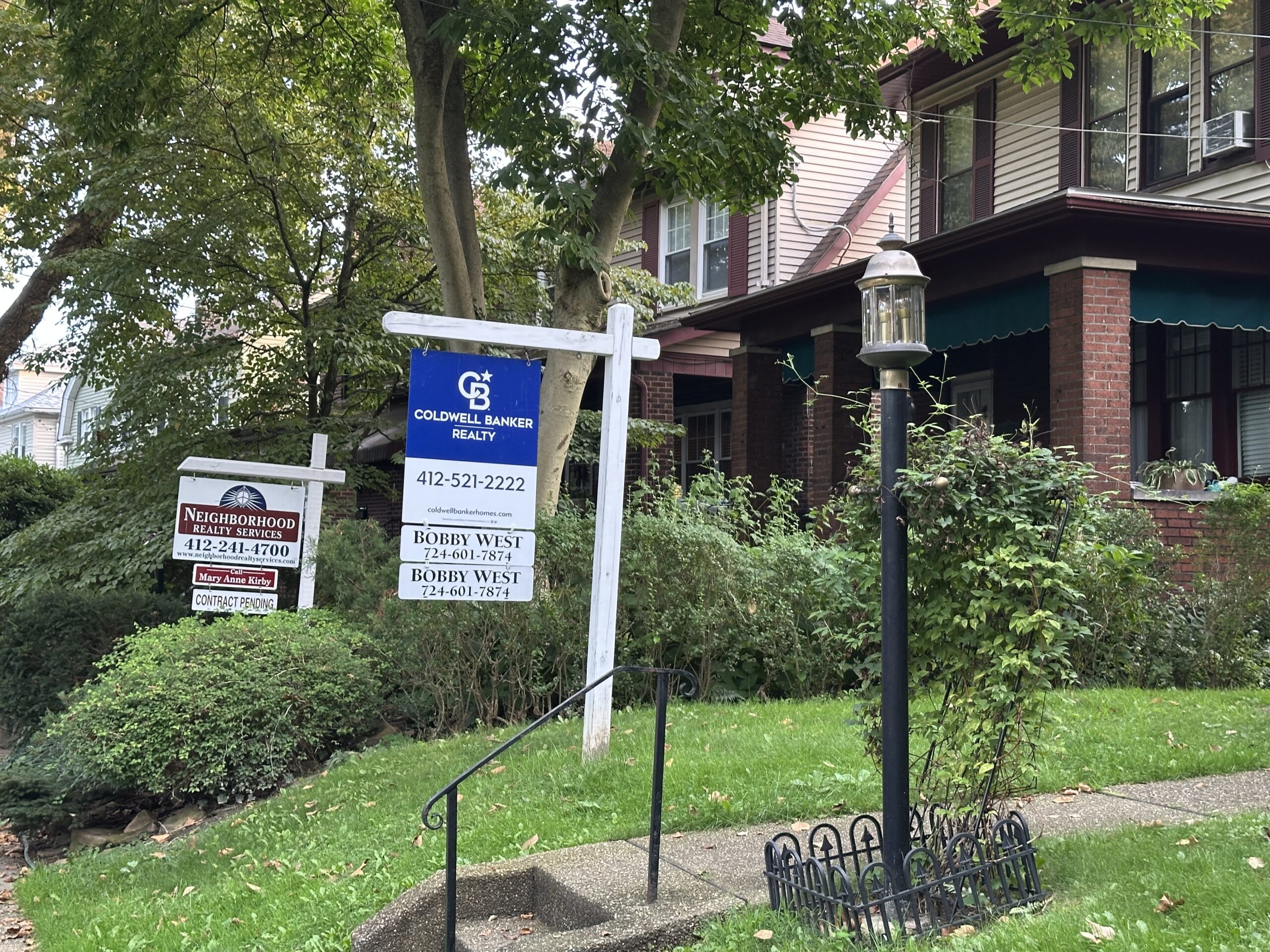 Brick house with two “For Sale” signs in front yard, surrounded by green lawn, trees, and a lamppost.