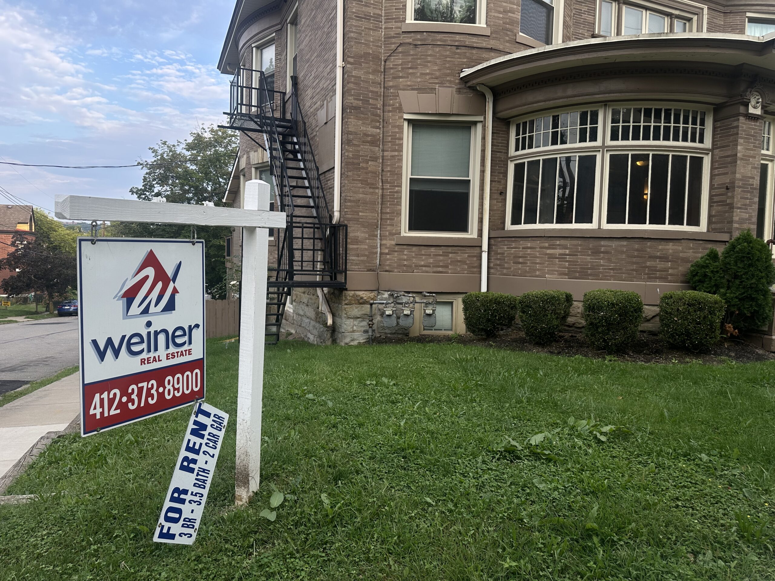 A “For Rent” sign from Weiner Real Estate stands on the lawn of a brick house with rounded windows and a black metal fire escape.