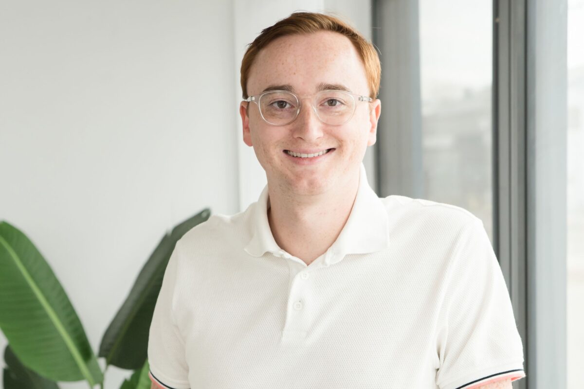 A person with short red hair and glasses, wearing a white polo shirt, stands indoors near a window and a green plant, smiling at the camera.