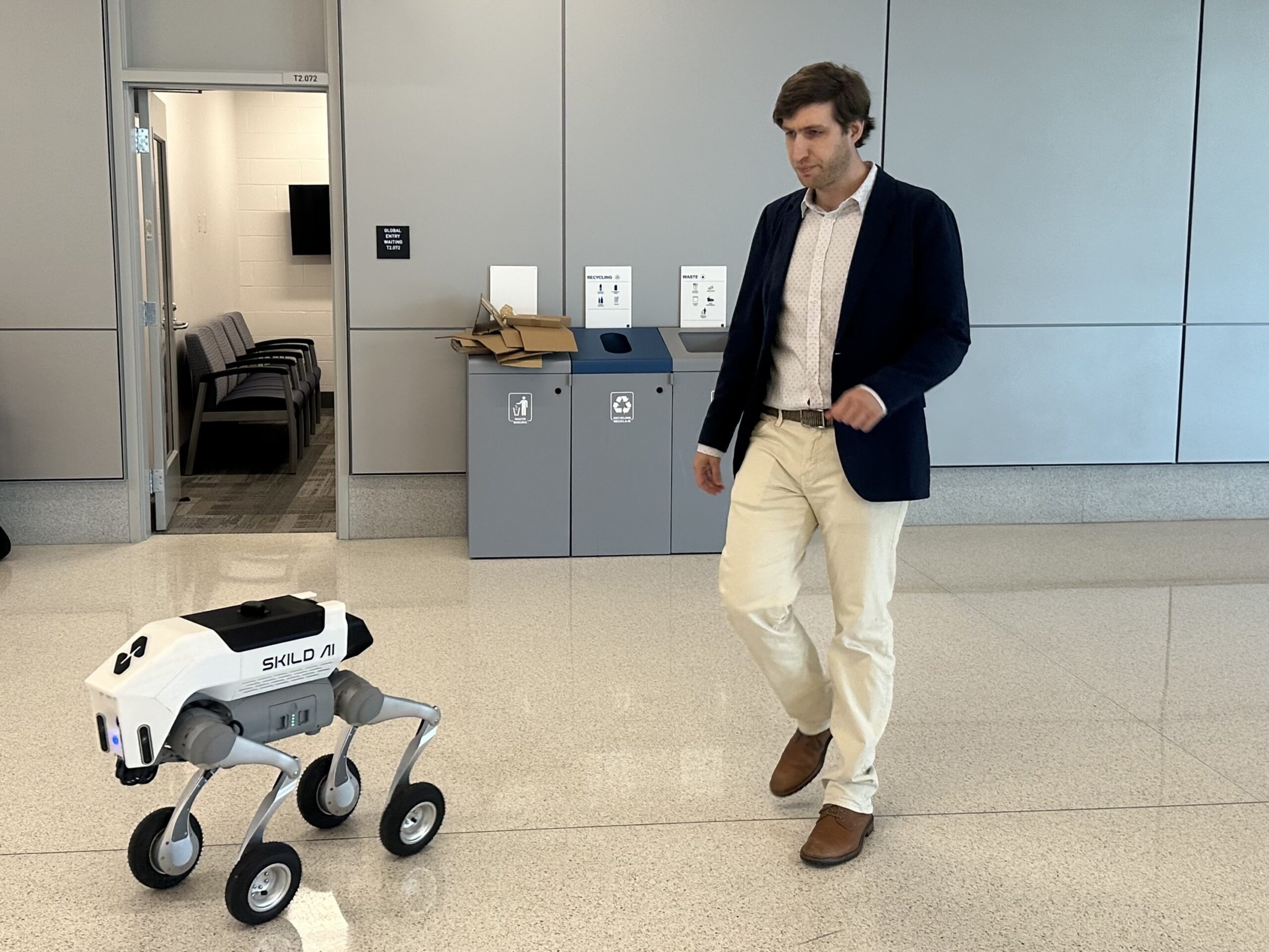 A man in business attire walks beside a small four-legged robot labeled “SKILLO AI” in a modern indoor space.