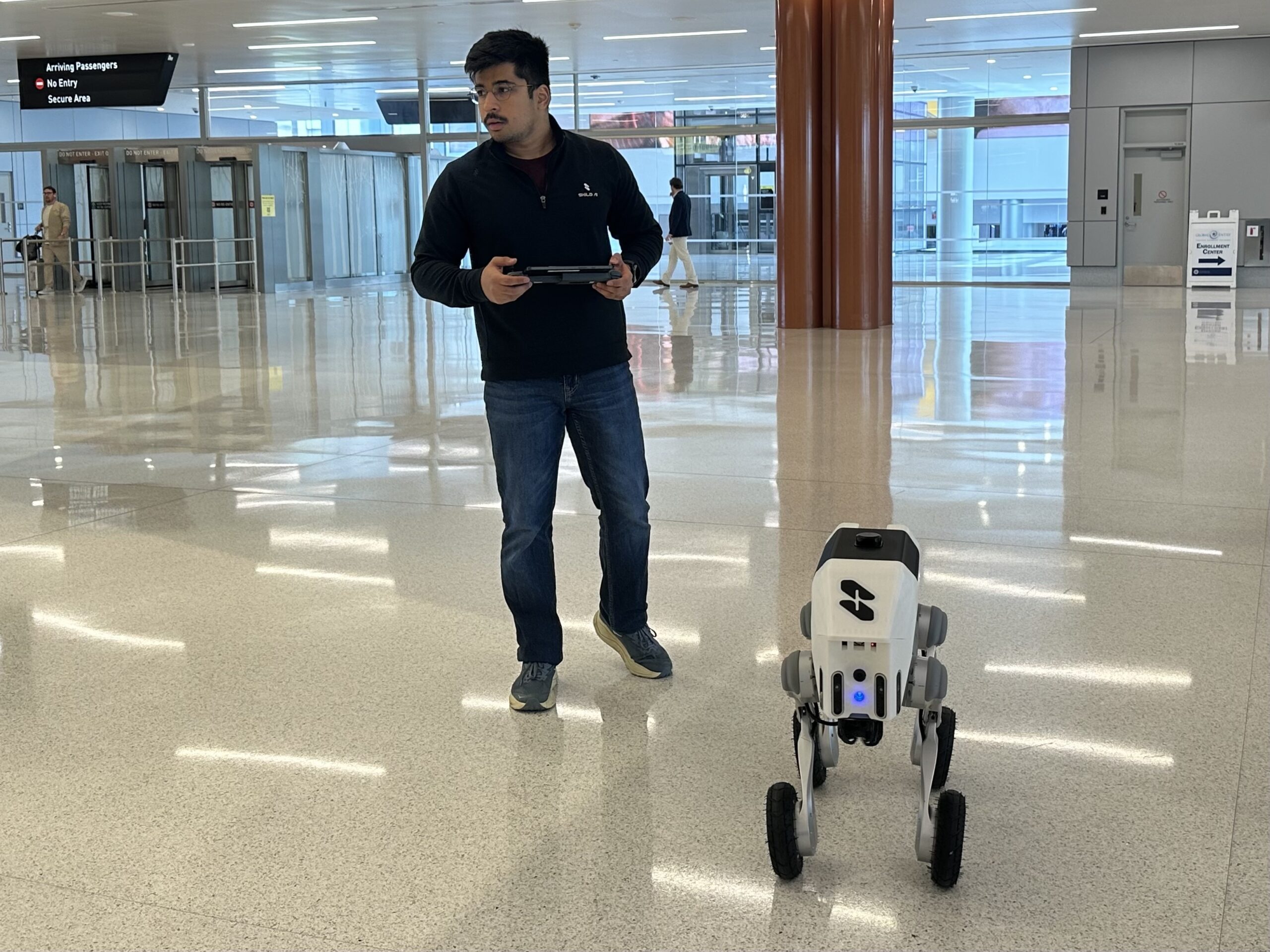 A person operates a four-legged robot with wheels inside a spacious, modern building with shiny floors and high ceilings.