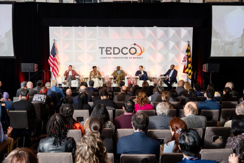 A panel of five people speaks on stage at a TEDCO event, facing an audience. The stage has US and Maryland flags and a large TEDCO sign behind the speakers.