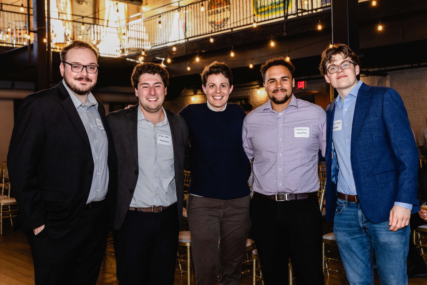 Five men stand together indoors, dressed in business casual attire, smiling at the camera. A modern loft setting with string lights is visible in the background.