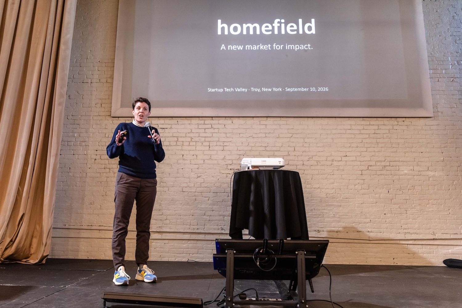 A person stands on stage giving a presentation titled “homefield: A new market for impact” at Startup Tech Valley, with a projected slide behind them.